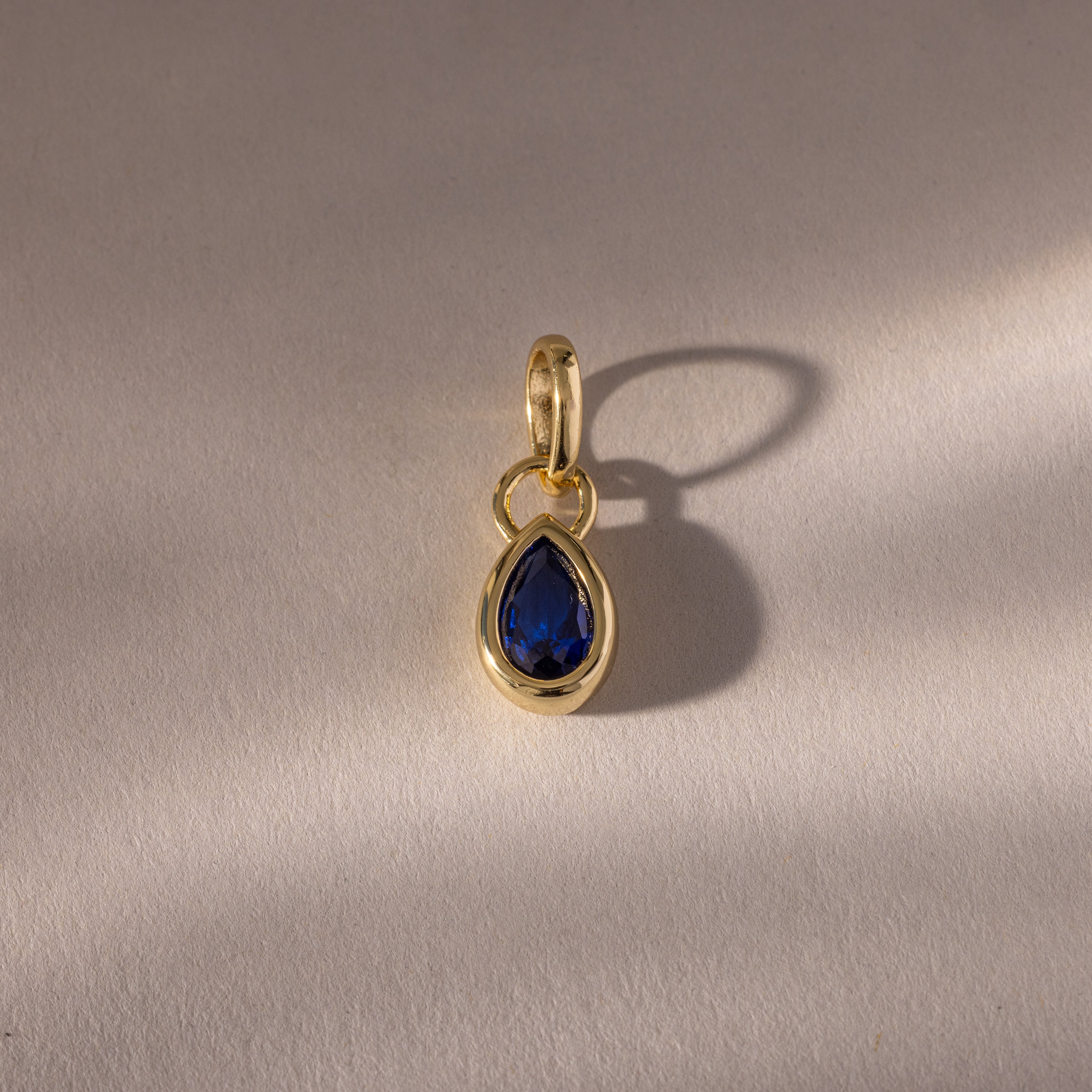 The Teardrop Sapphire Charm is a gold pendant with a teardrop-shaped sapphire in a bezel setting, featuring a blue gemstone that casts a shadow on a light textured surface.
