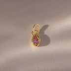Teardrop Pink Tourmaline Charm: A gold pendant featuring a teardrop-shaped pink tourmaline in a gold bezel setting, displayed on a light textured surface. A timeless piece of gemstone jewelry, perfect as a birthstone charm.