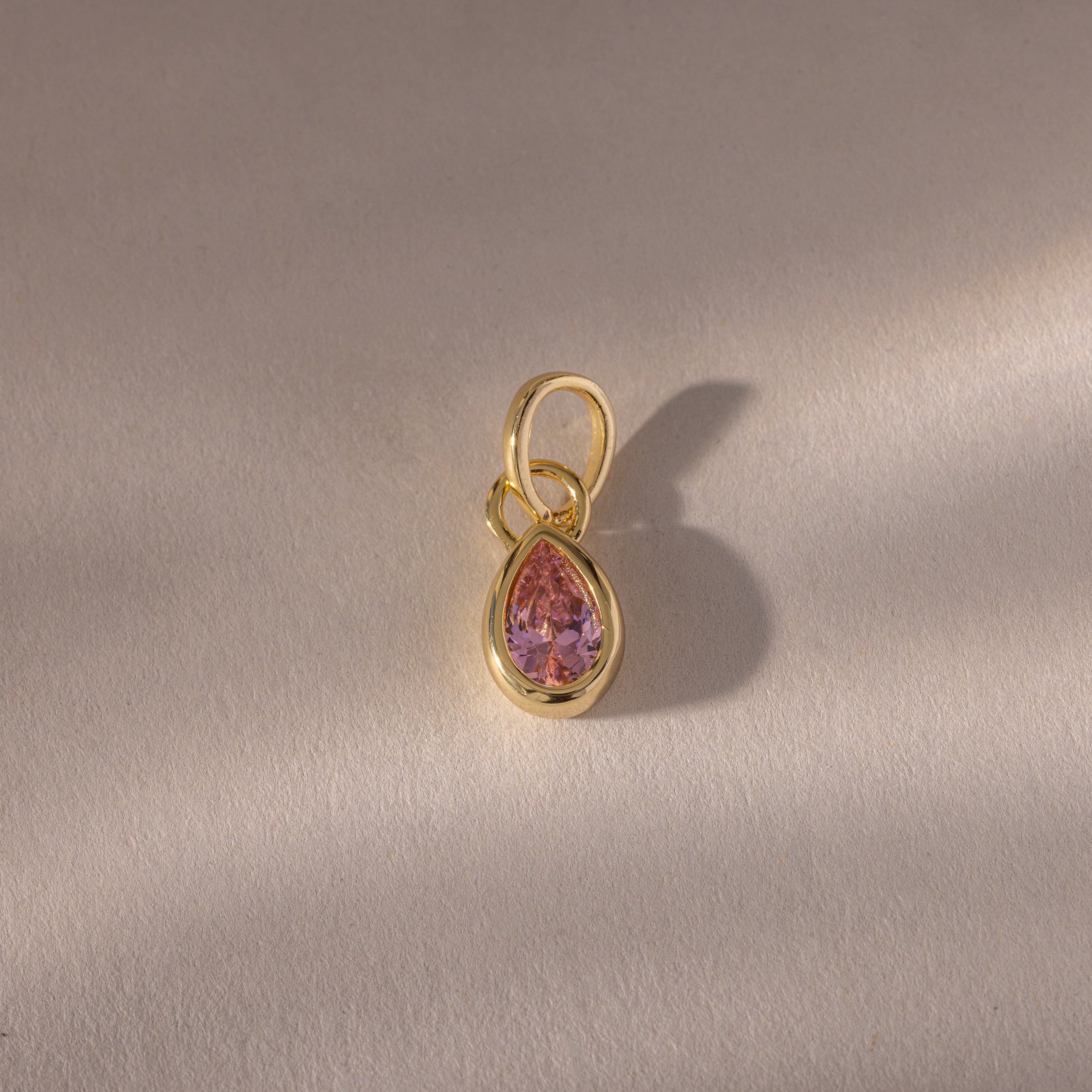 Teardrop Pink Tourmaline Charm: A gold pendant featuring a teardrop-shaped pink tourmaline in a gold bezel setting, displayed on a light textured surface. A timeless piece of gemstone jewelry, perfect as a birthstone charm.