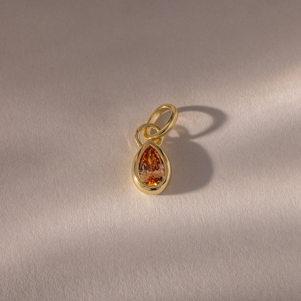The Teardrop Topaz Charm features an orange gemstone in a gold bezel setting, casting a shadow on a textured light surface.