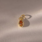 The Teardrop Topaz Charm features an orange gemstone in a gold bezel setting, casting a shadow on a textured light surface.