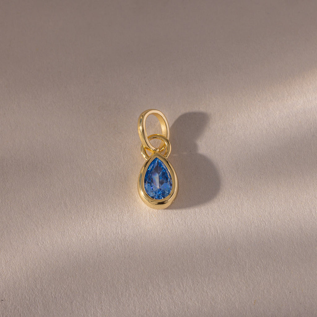 The Teardrop Blue Zircon Charm features a teardrop-shaped blue zircon in a gold bezel setting, displayed on a softly textured surface—an elegant gemstone jewelry piece.