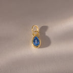 The Teardrop Blue Zircon Charm features a teardrop-shaped blue zircon in a gold bezel setting, displayed on a softly textured surface—an elegant gemstone jewelry piece.