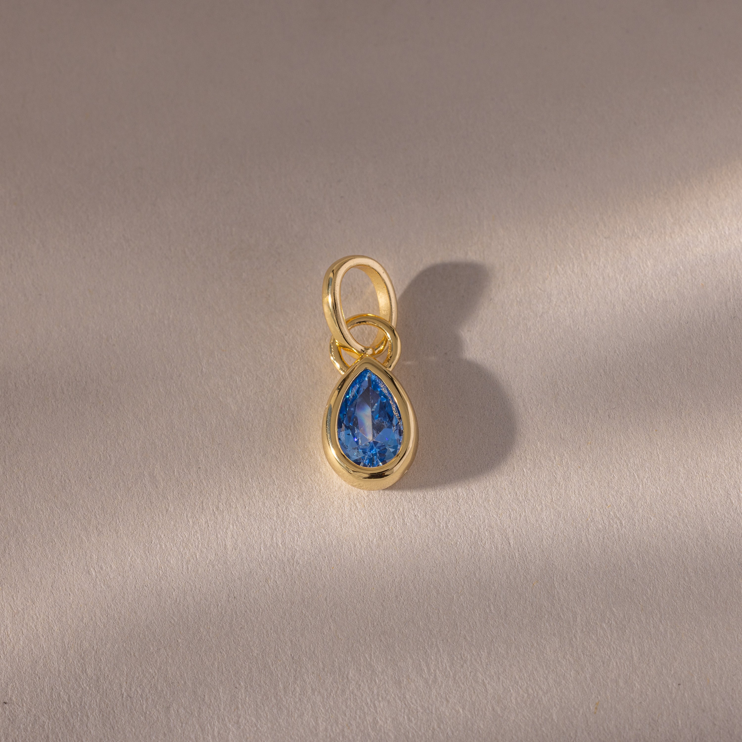 The Teardrop Blue Zircon Charm features a teardrop-shaped blue zircon in a gold bezel setting, displayed on a softly textured surface—an elegant gemstone jewelry piece.