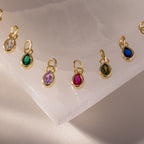 Teardrop Birthstone Charms, each set in a delicate gold bezel, are arranged in a row on white marble, showcasing classic colorful gemstone style.