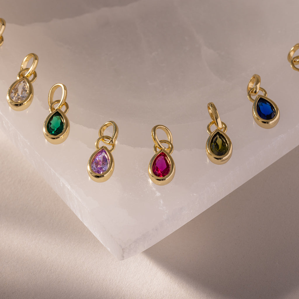 Teardrop Birthstone Charm pendants in gold bezel settings, elegantly displayed on white marble.
