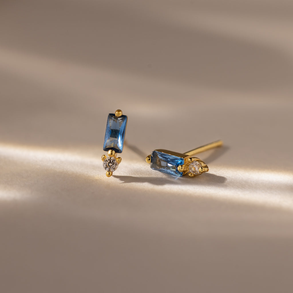 Baguette Blue Zircon Drop Studs showcase rectangular blue zircon stones and teardrop clear gems, creating a modern, elegant earrings look on a neutral surface.