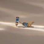 Baguette Blue Zircon Drop Studs showcase rectangular blue zircon stones and teardrop clear gems, creating a modern, elegant earrings look on a neutral surface.