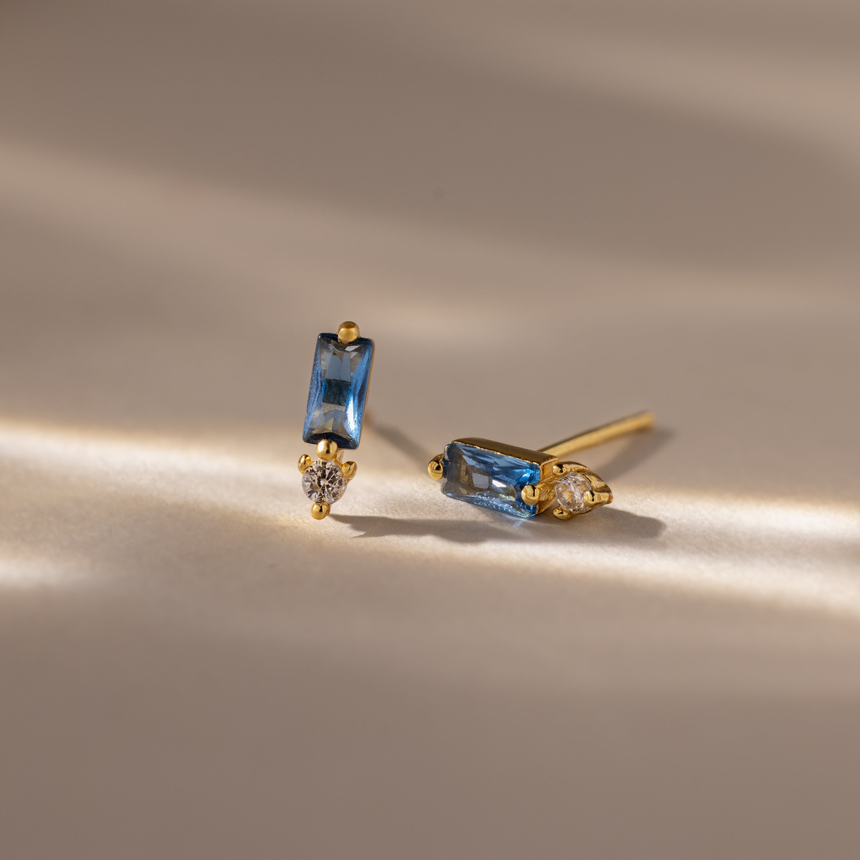 Baguette Blue Zircon Drop Studs showcase rectangular blue zircon stones and teardrop clear gems, creating a modern, elegant earrings look on a neutral surface.