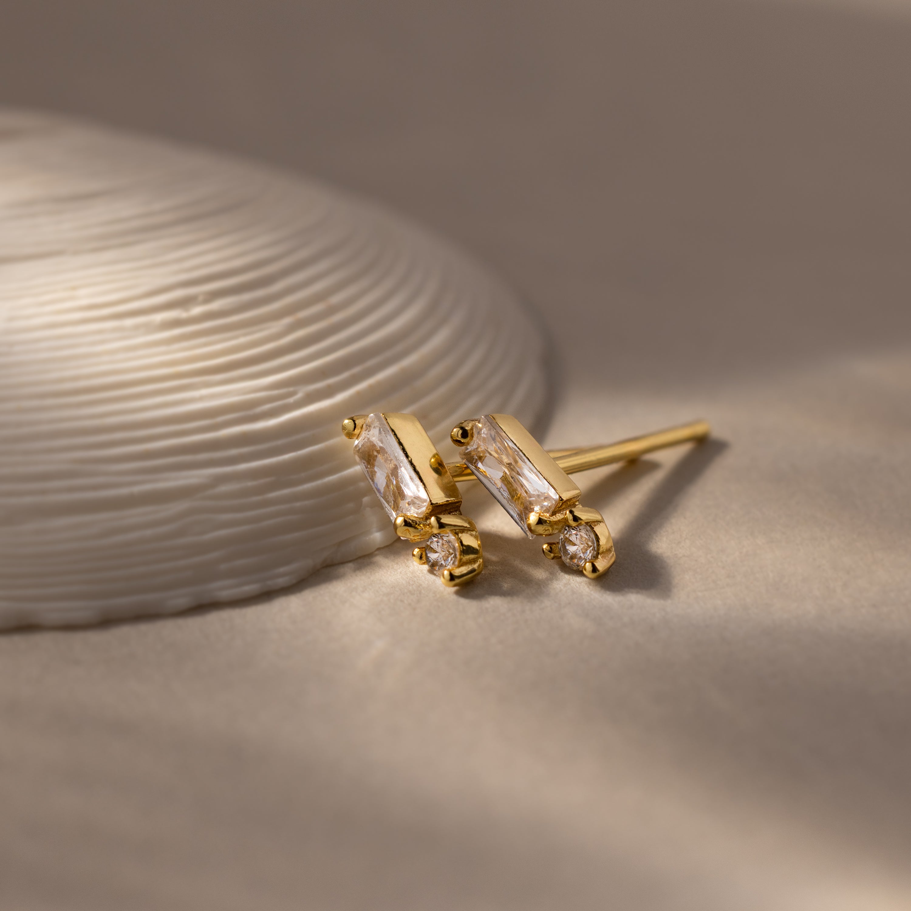 Baguette Birthstone Drop Studs showcase rectangular stones with small round gems, offering a modern jewelry look and displayed next to a textured white shell.