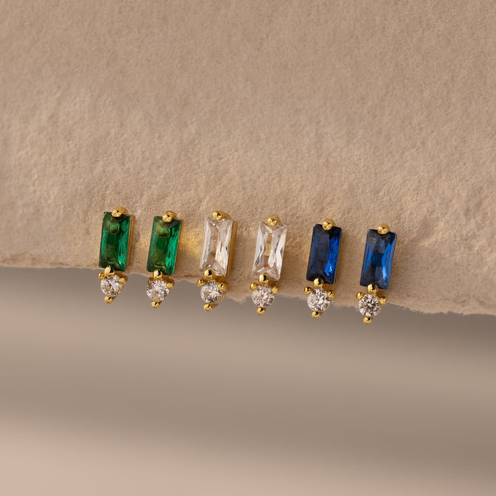 Three pairs of Baguette Birthstone Drop Studs: gold earrings with green, clear, and blue baguette stones, each accented by a tiny diamond.