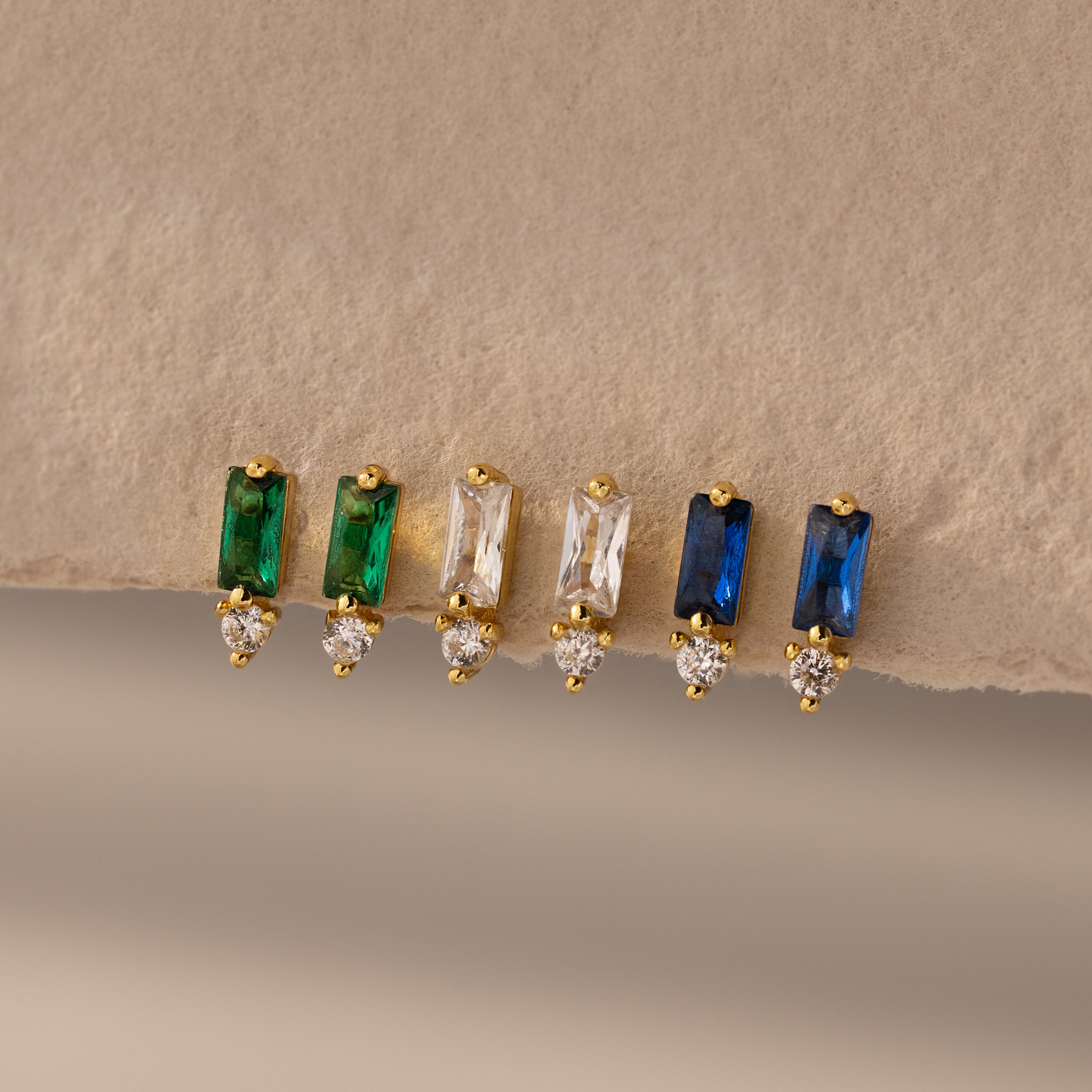 Three pairs of Baguette Birthstone Drop Studs: gold earrings with green, clear, and blue baguette stones, each accented by a tiny diamond.