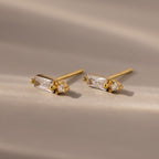 The Baguette Diamond Drop Studs feature gold settings with rectangular and round clear gemstones, offering a modern, elegant look displayed on a softly lit neutral surface.
