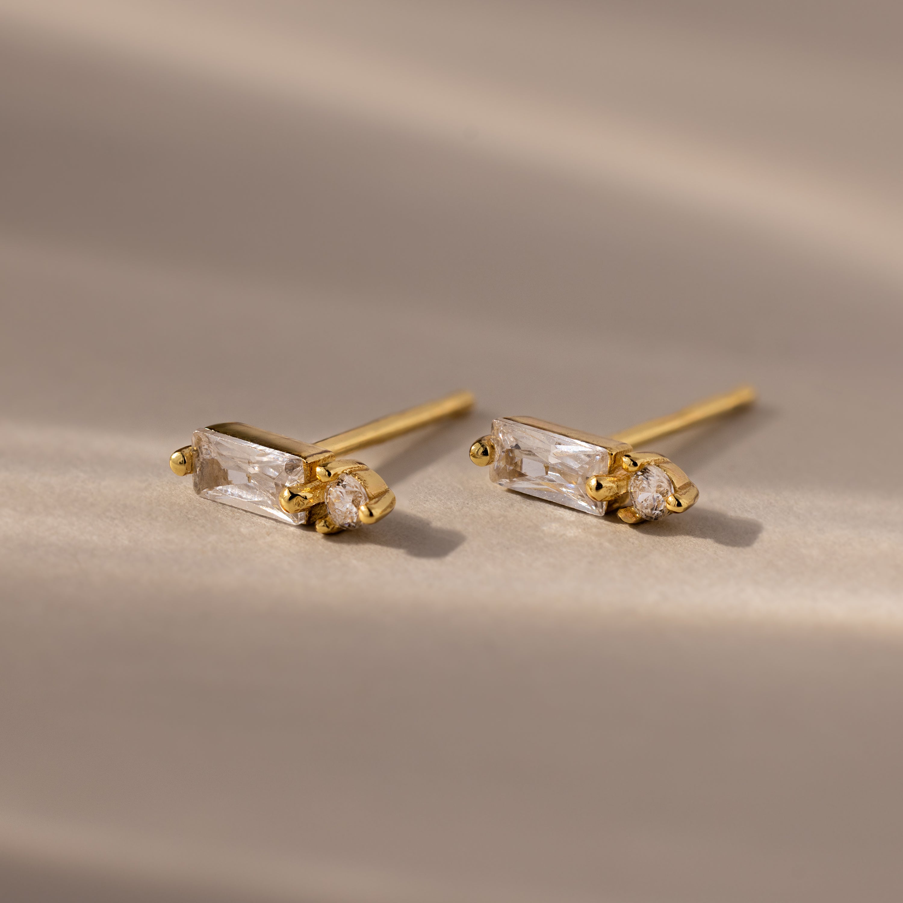 The Baguette Diamond Drop Studs feature gold settings with rectangular and round clear gemstones, offering a modern, elegant look displayed on a softly lit neutral surface.