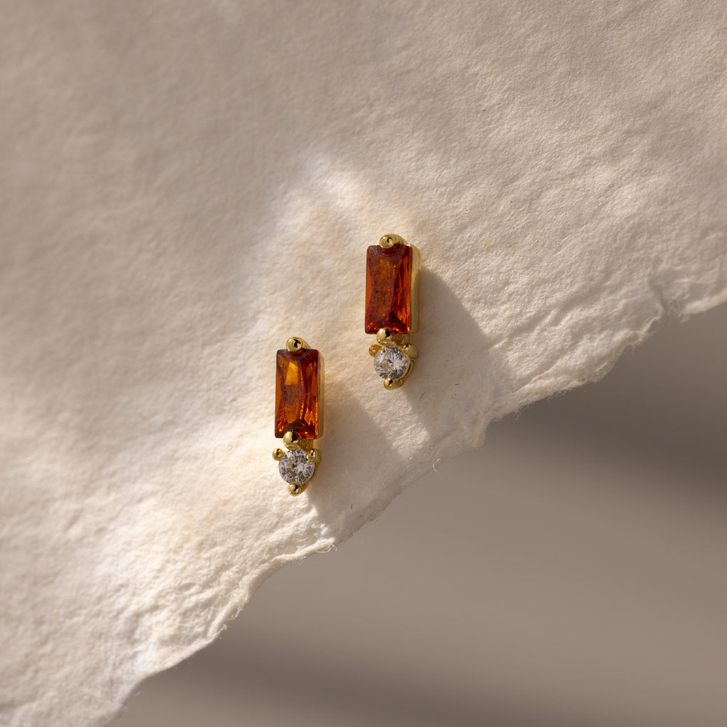 Baguette Garnet Drop Studs featuring rectangular garnet stones and round clear stones are displayed on a textured cream paper background—a perfect touch of modern jewelry elegance.