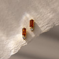 Baguette Garnet Drop Studs featuring rectangular garnet stones and round clear stones are displayed on a textured cream paper background—a perfect touch of modern jewelry elegance.