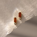 Baguette Garnet Drop Studs featuring rectangular garnet stones and round clear stones are displayed on a textured cream paper background—a perfect touch of modern jewelry elegance.