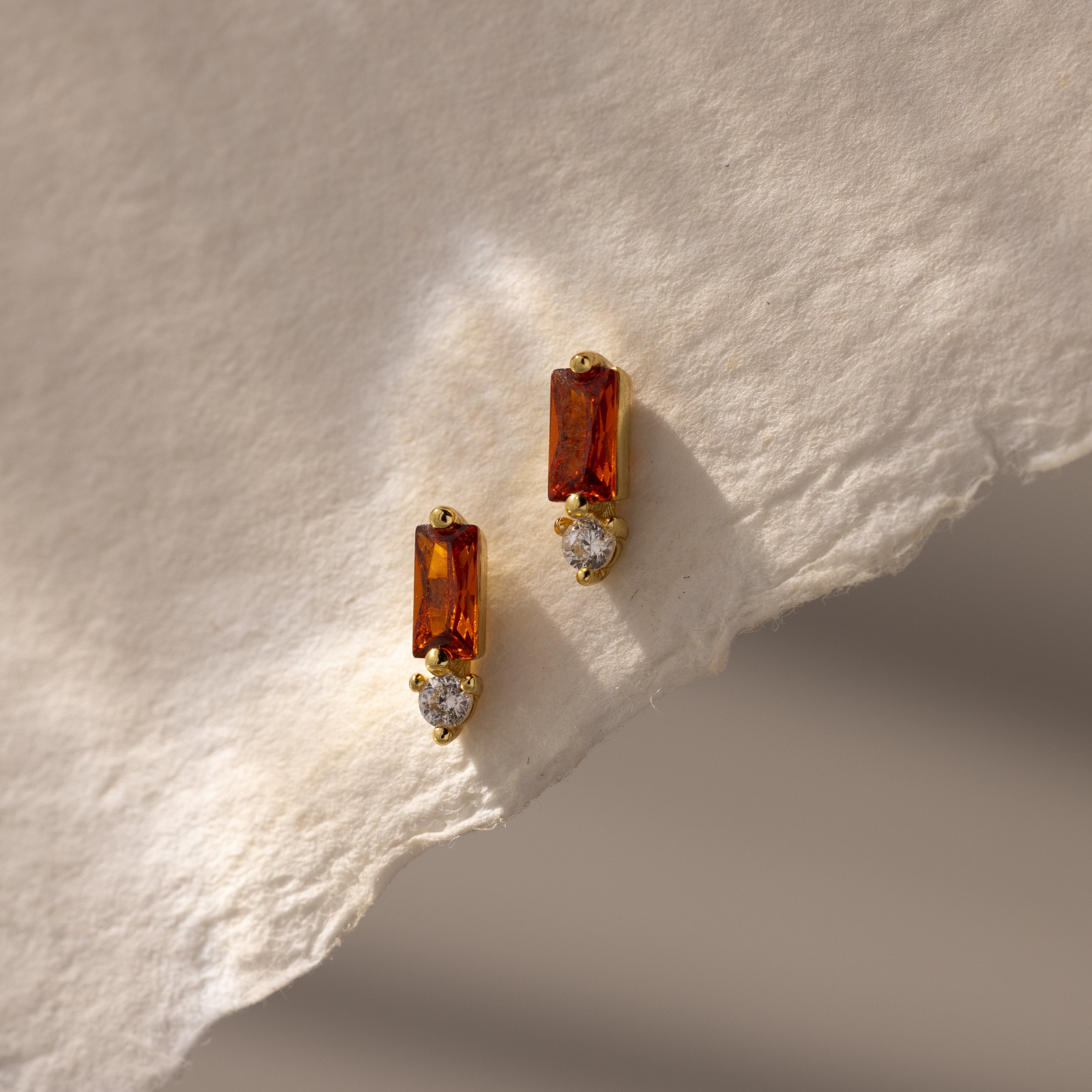 Baguette Garnet Drop Studs featuring rectangular garnet stones and round clear stones are displayed on a textured cream paper background—a perfect touch of modern jewelry elegance.