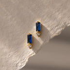 Gold earrings with rectangular blue stones and small round white gems, the Baguette Sapphire Drop Studs are a modern, elegant jewelry piece, beautifully displayed on a textured off-white paper background.