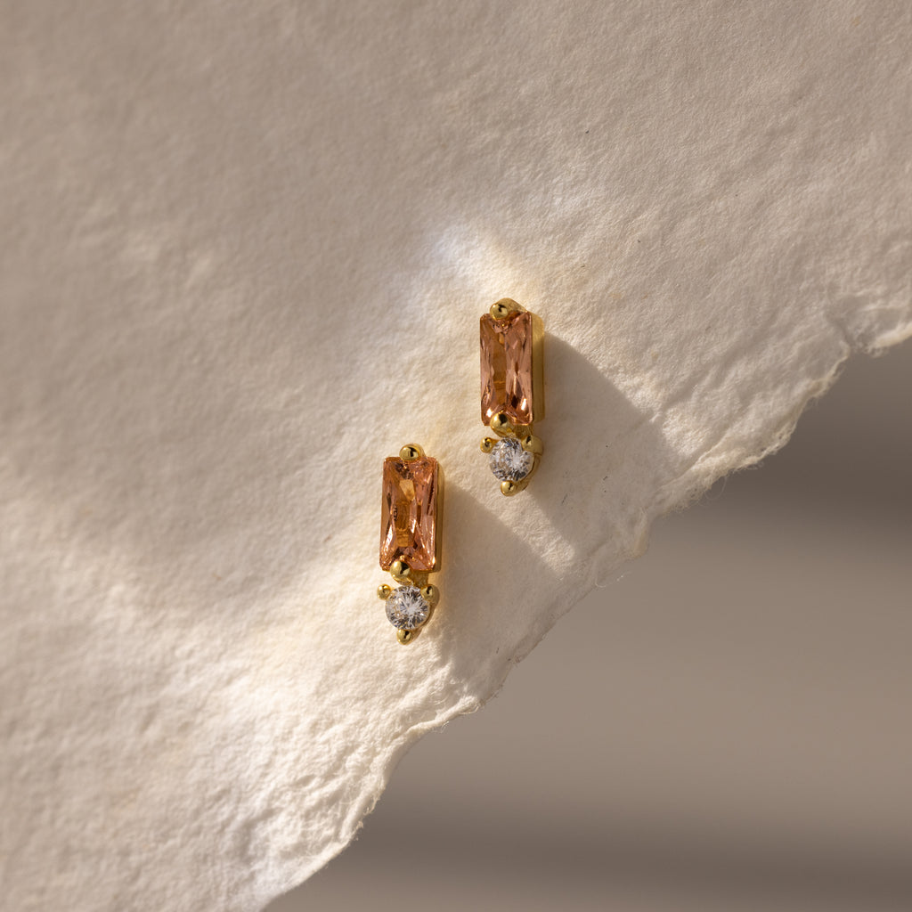 Baguette Topaz Drop Studs: gold earrings with rectangular peach topaz and small round diamonds, styled on textured paper. The perfect touch of elegant birthstone jewelry.