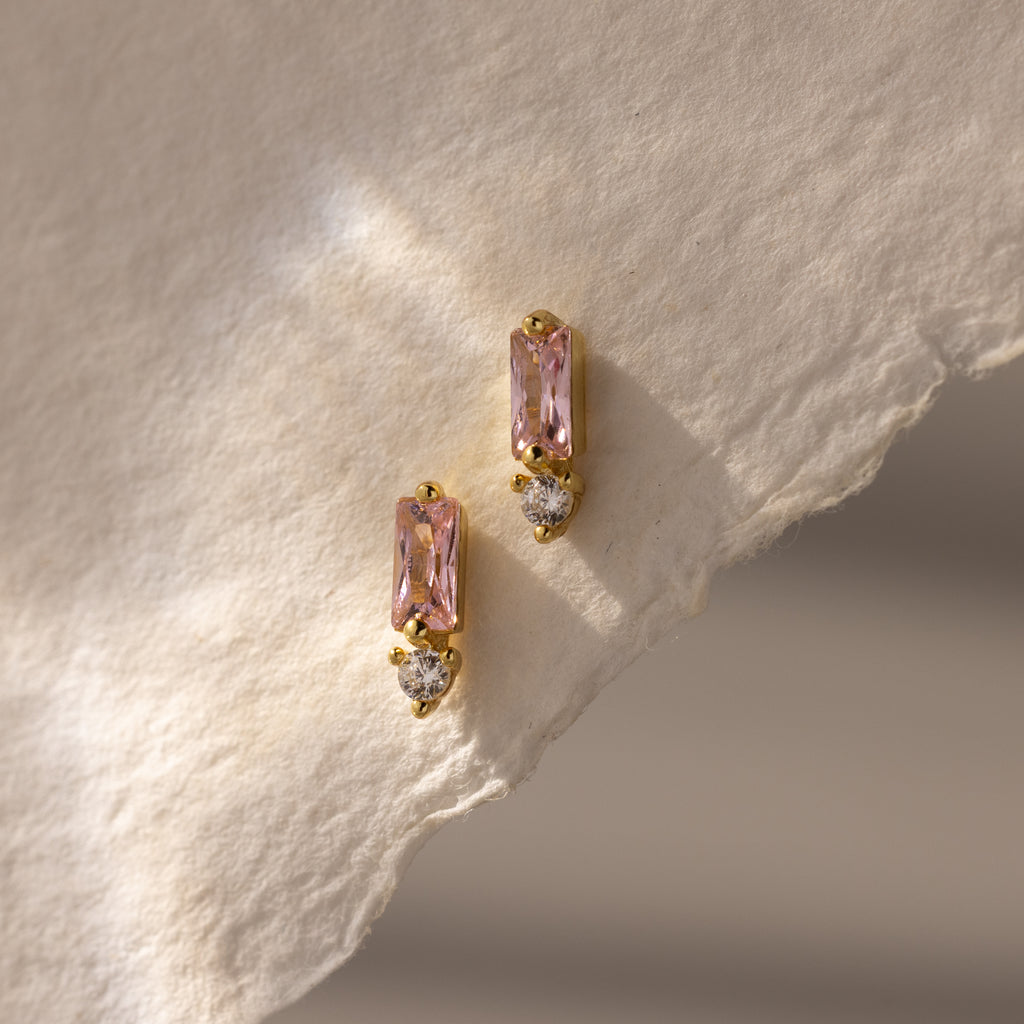 Baguette Pink Tourmaline Drop Studs: Modern gold earrings with pink rectangular tourmaline and small clear stones, displayed on textured paper, bringing a fresh update to classic birthstone earrings.