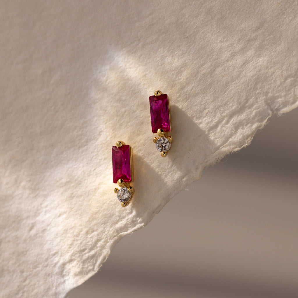 Baguette Ruby Drop Studs feature rectangular red stones paired with small round clear stones, displayed on textured beige paper—ideal for fans of modern jewelry.