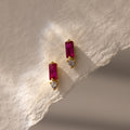 Baguette Ruby Drop Studs feature rectangular red stones paired with small round clear stones, displayed on textured beige paper—ideal for fans of modern jewelry.