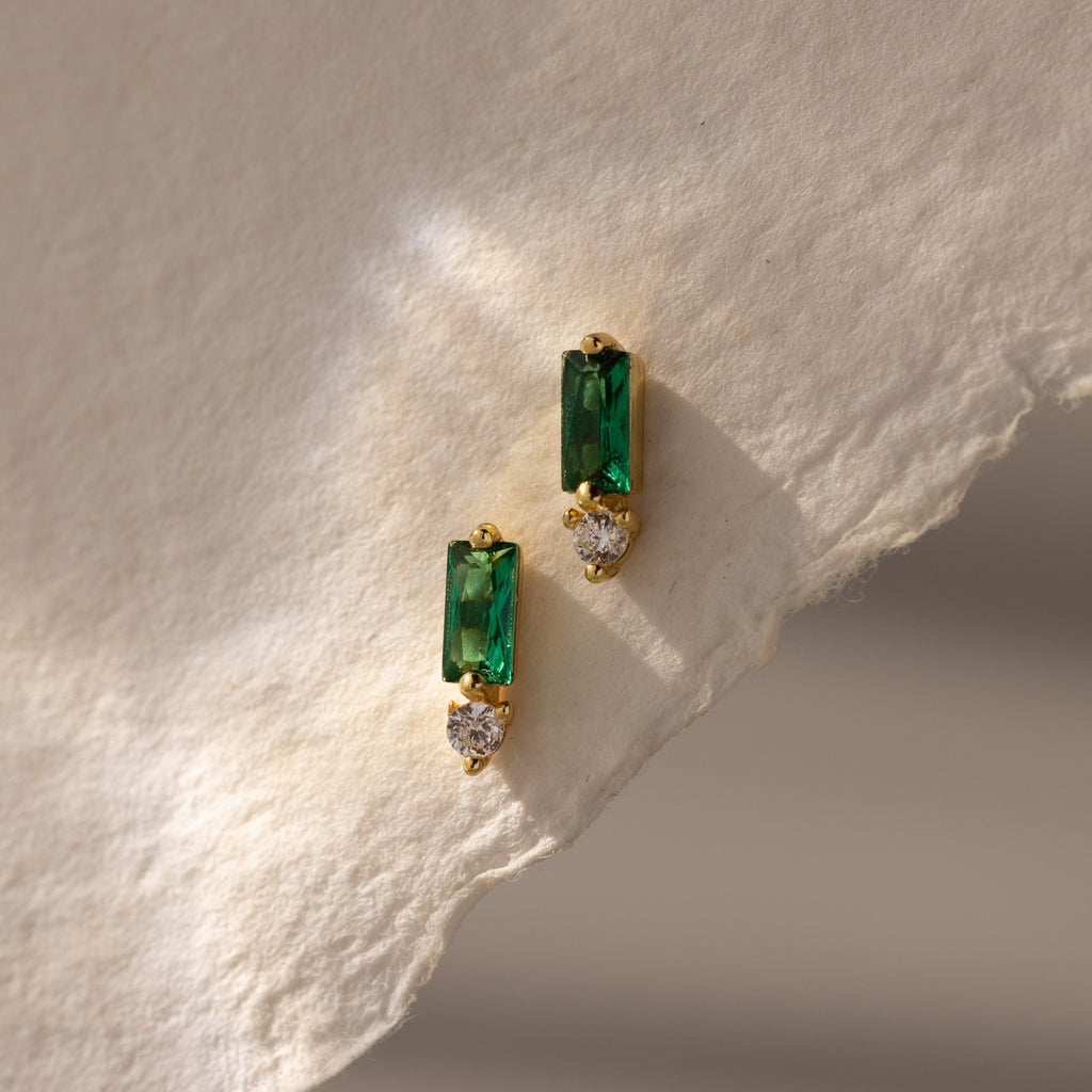 Baguette Emerald Drop Studs feature green rectangular stones and small round clear gems, displayed on textured, light paper—ideal for fans of modern, elegant jewelry.