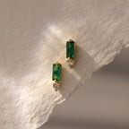 Baguette Emerald Drop Studs feature green rectangular stones and small round clear gems, displayed on textured, light paper—ideal for fans of modern, elegant jewelry.