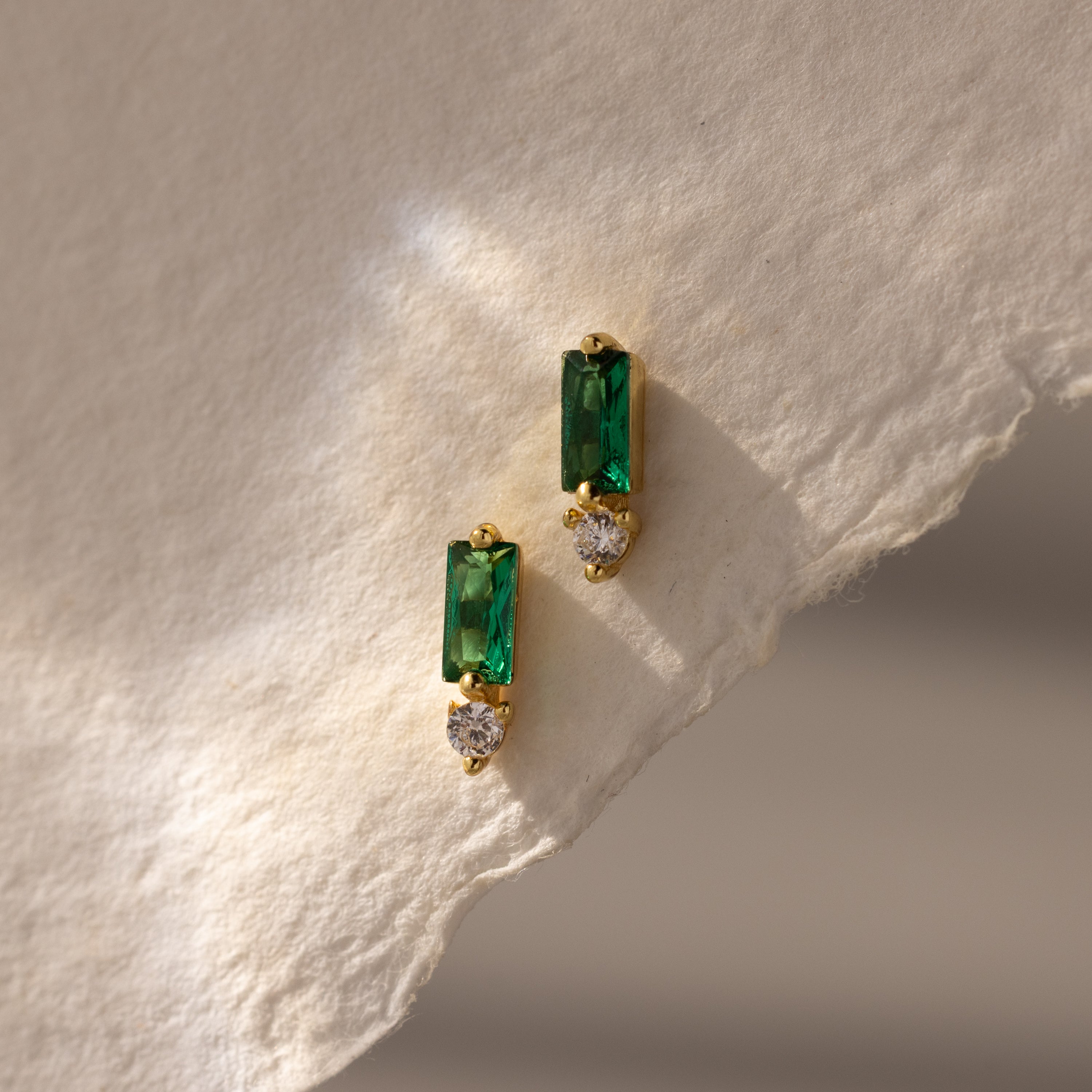 Baguette Emerald Drop Studs feature green rectangular stones and small round clear gems, displayed on textured, light paper—ideal for fans of modern, elegant jewelry.