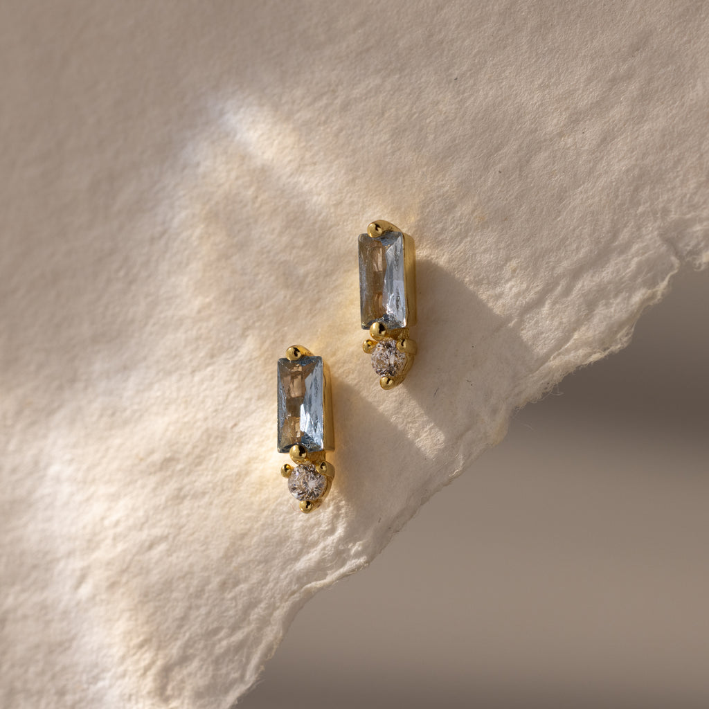 Modern elegant jewelry: these Baguette Aquamarine Drop Studs feature gold earrings with rectangular aquamarine stones and small round gems, beautifully displayed on textured cream paper.
