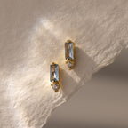 Modern elegant jewelry: these Baguette Aquamarine Drop Studs feature gold earrings with rectangular aquamarine stones and small round gems, beautifully displayed on textured cream paper.