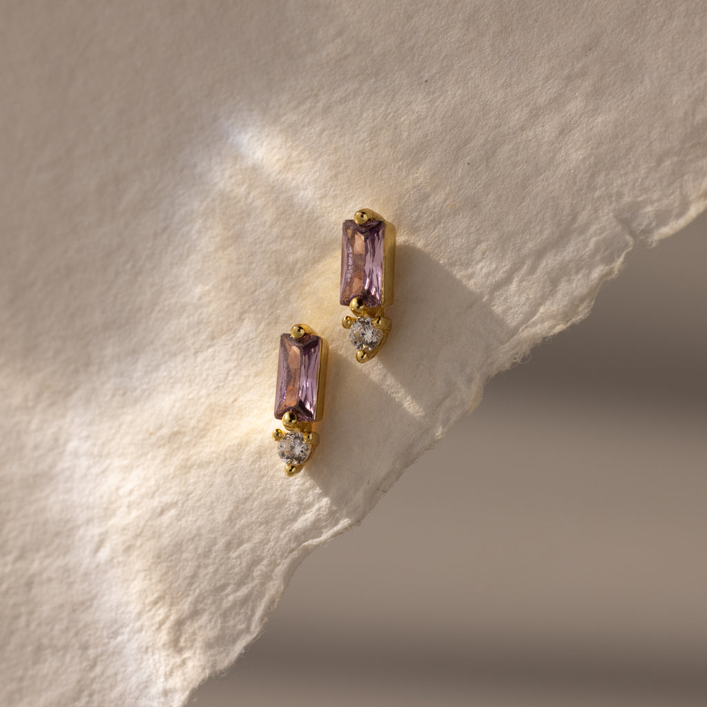 Two Baguette Alexandrite Drop Studs with purple rectangular stones and small clear gems rest on textured cream paper, showcasing modern, elegant earrings perfect for any occasion.