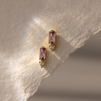 Two Baguette Alexandrite Drop Studs with purple rectangular stones and small clear gems rest on textured cream paper, showcasing modern, elegant earrings perfect for any occasion.