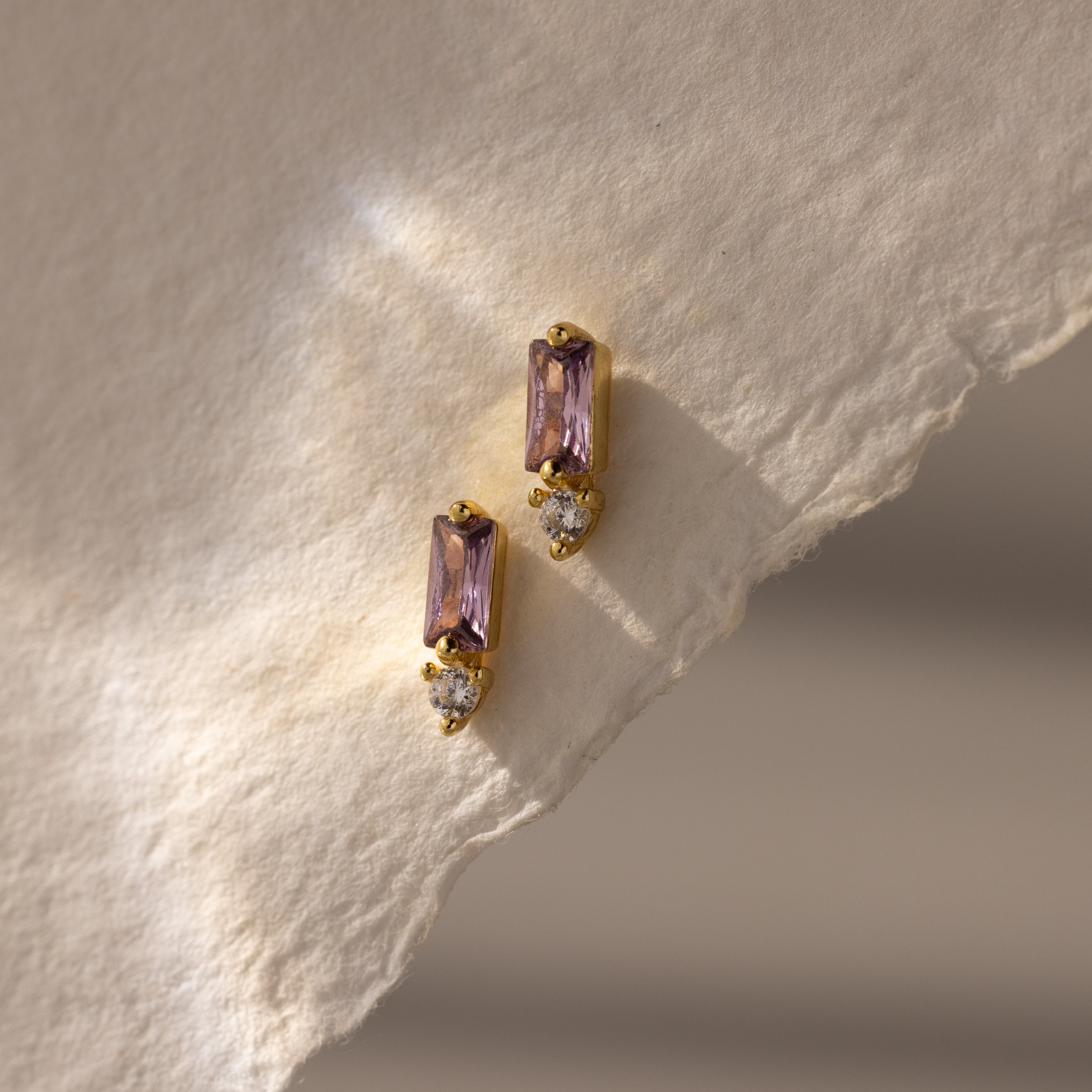 Two Baguette Alexandrite Drop Studs with purple rectangular stones and small clear gems rest on textured cream paper, showcasing modern, elegant earrings perfect for any occasion.