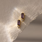 Baguette Amethyst Drop Studs featuring rectangular purple stones and clear gems, displayed on textured off-white paper—an elegant and modern interpretation of birthstone earrings.