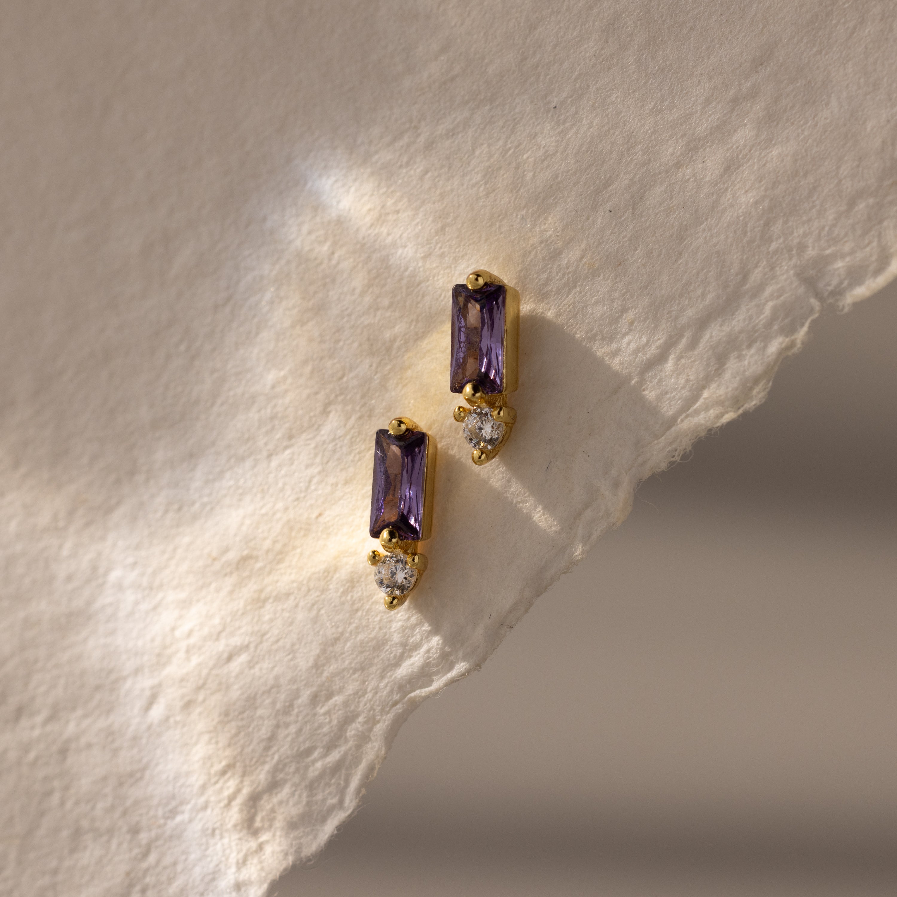 Baguette Amethyst Drop Studs featuring rectangular purple stones and clear gems, displayed on textured off-white paper—an elegant and modern interpretation of birthstone earrings.