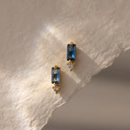 Two Baguette Blue Zircon Drop Studs with blue rectangular stones and small clear gems, displayed on textured beige paper—modern, elegant earrings perfect for any occasion.