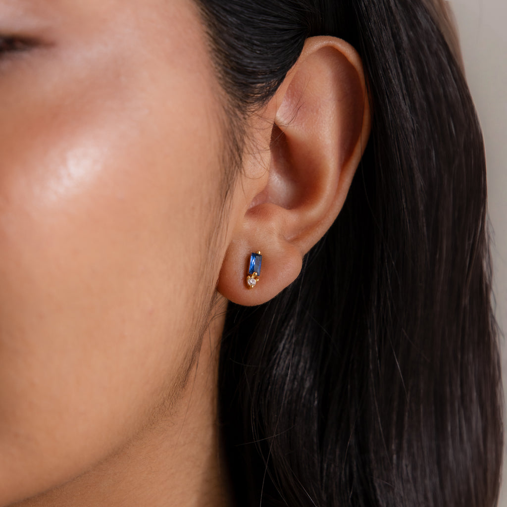 Close-up of a woman's ear with straight dark hair, wearing Baguette Blue Zircon Drop Studs—perfect modern elegant earrings for those who love blue and gold or birthstone jewelry.