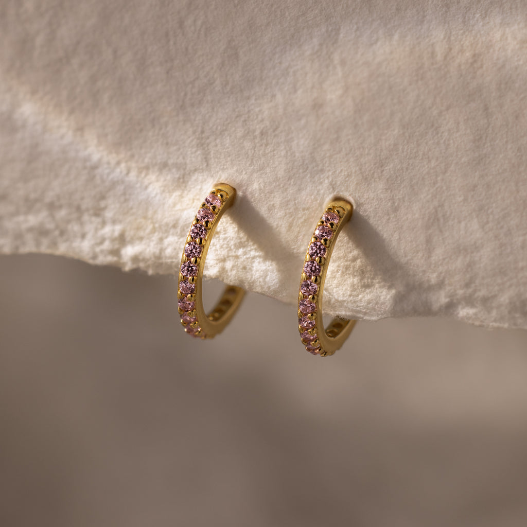 Pink Tourmaline Clicker Huggies—gold hoop earrings adorned with small pink tourmaline gemstones, ideal for daily wear, shown resting on a soft textured fabric.