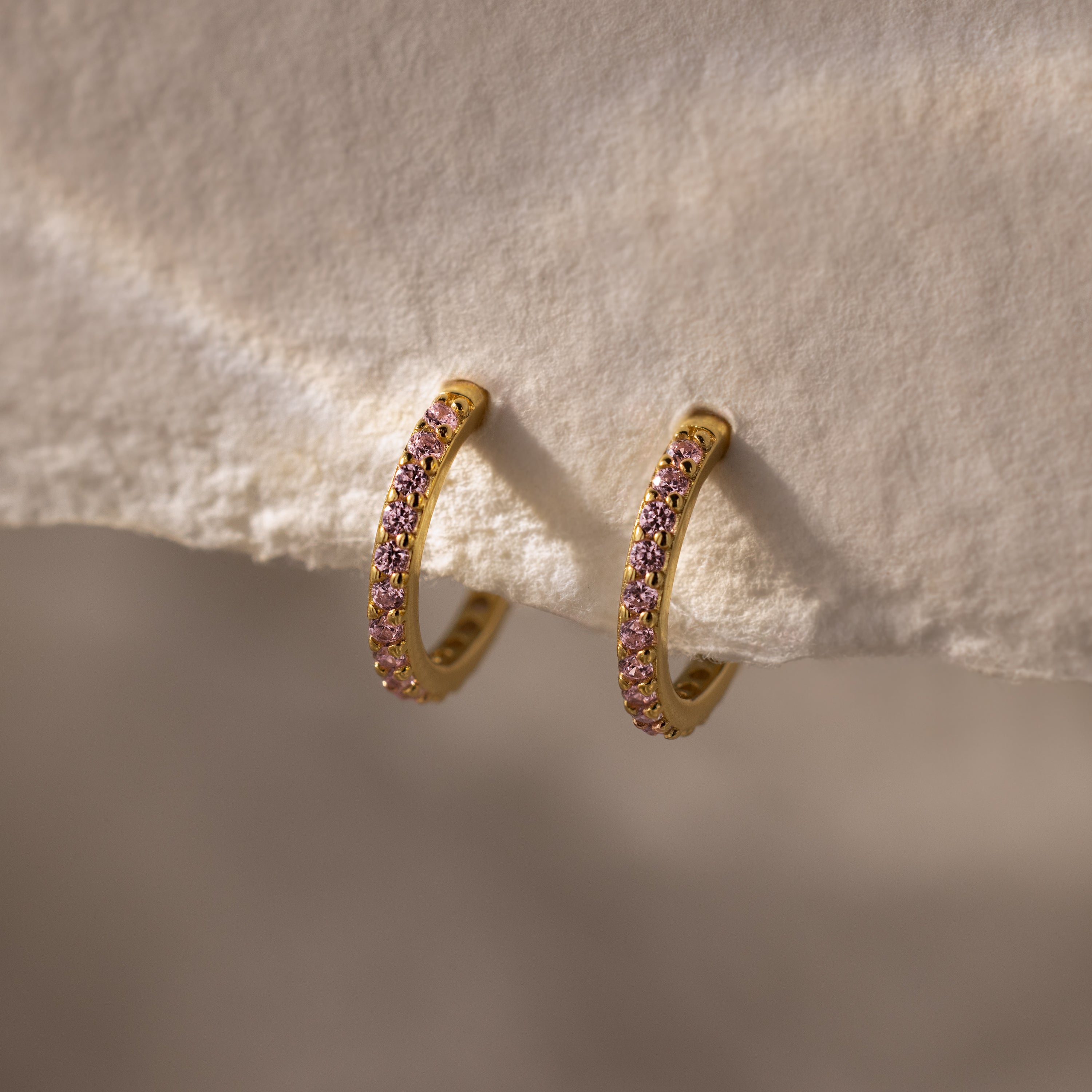 Pink Tourmaline Clicker Huggies—gold hoop earrings adorned with small pink tourmaline gemstones, ideal for daily wear, shown resting on a soft textured fabric.