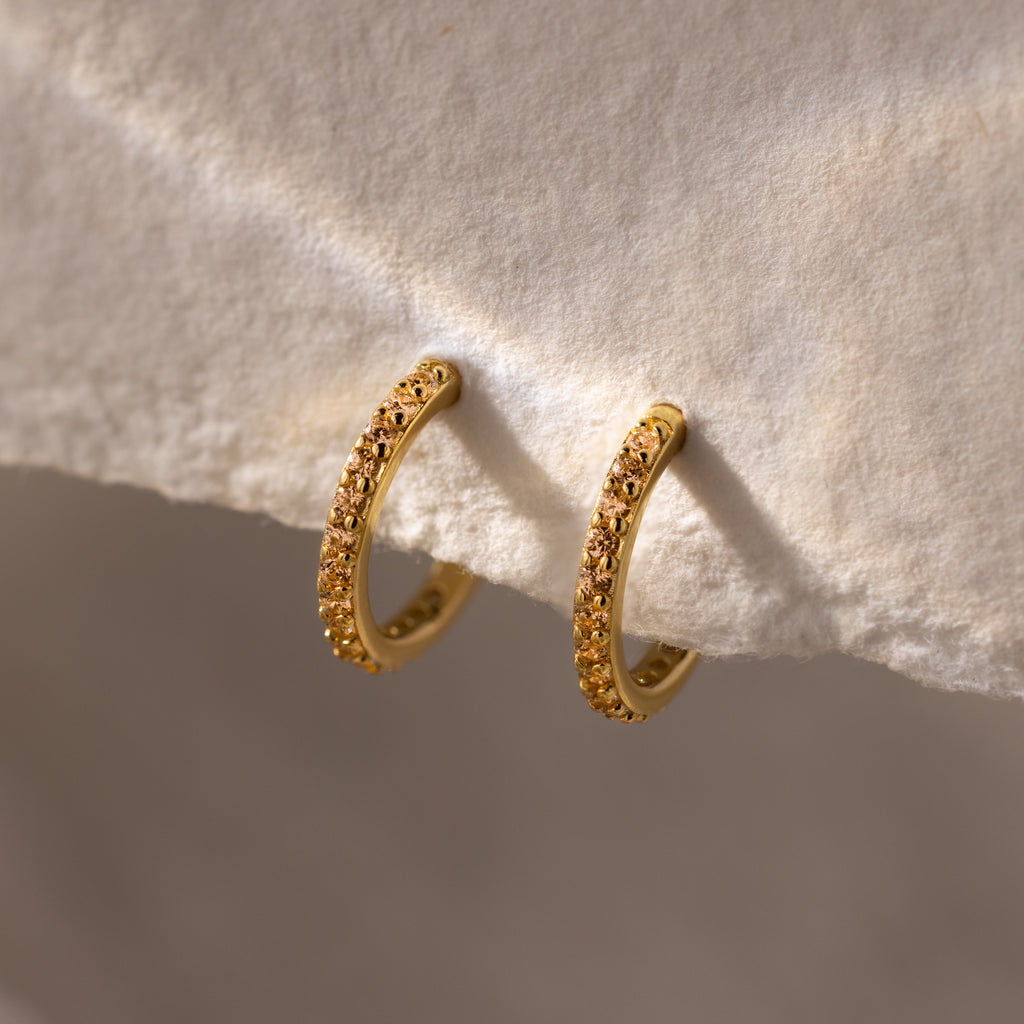 Topaz Clicker Huggies feature gold hoops with small sparkling stones, displayed on a textured beige surface—perfect for adding subtle sparkle to your everyday look.