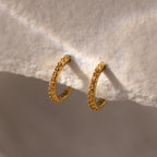 Topaz Clicker Huggies feature gold hoops with small sparkling stones, displayed on a textured beige surface—perfect for adding subtle sparkle to your everyday look.