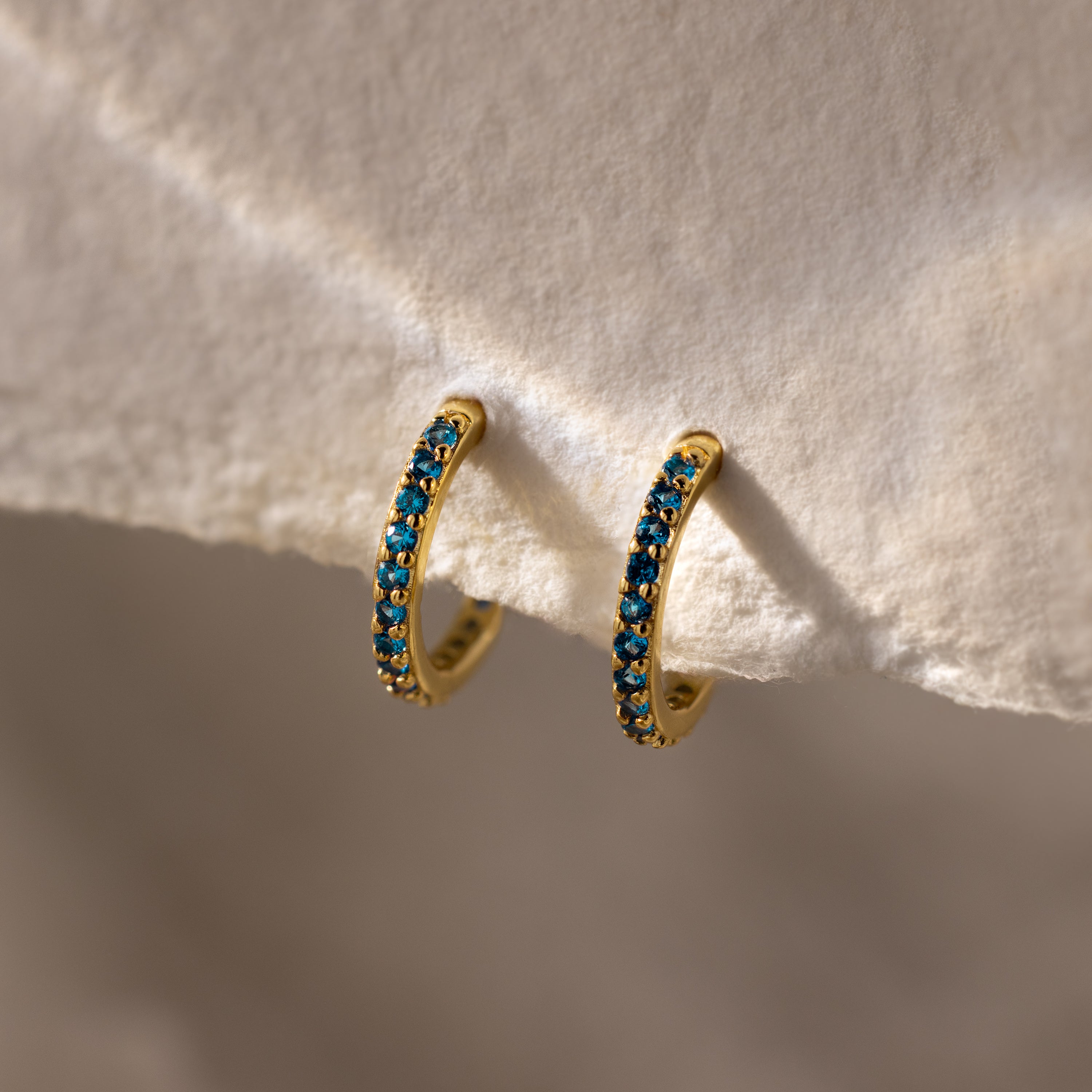 Blue Zircon Clicker Huggies—gold hoop earrings set with small blue zircon stones, shown on a textured cream background. These birthstone huggies are the perfect everyday accessory for a touch of elegant sparkle.