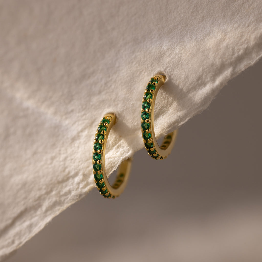 Emerald Clicker Huggies—gold hoop earrings featuring green gemstones, perfect as birthstone earrings for everyday wear, shown on a textured white surface.