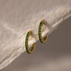 Emerald Clicker Huggies—gold hoop earrings featuring green gemstones, perfect as birthstone earrings for everyday wear, shown on a textured white surface.