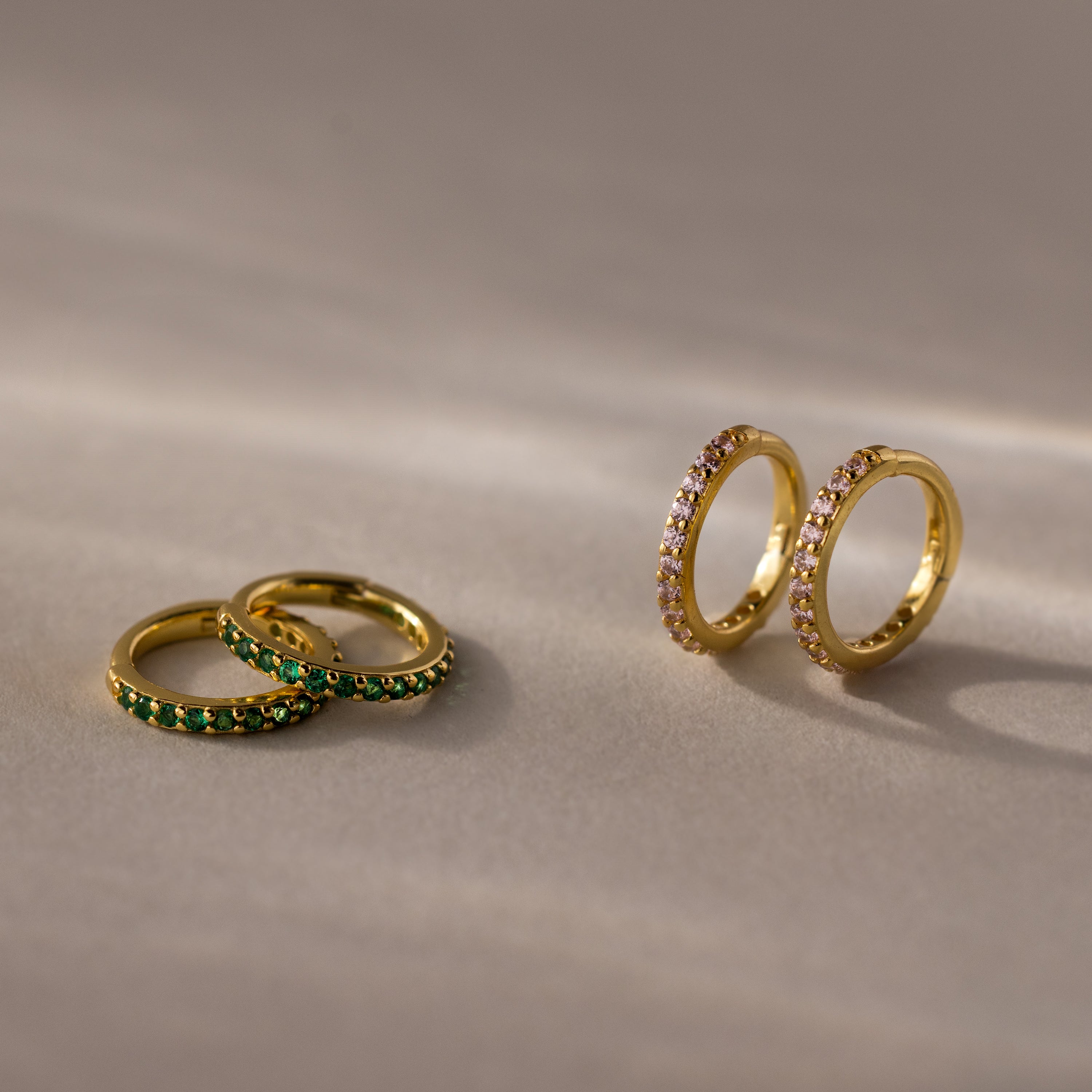 Two pairs of gold hoop earrings, including Pink Tourmaline Clicker Huggies, rest on a beige surface—ideal as birthstone earrings or for everyday huggie style.