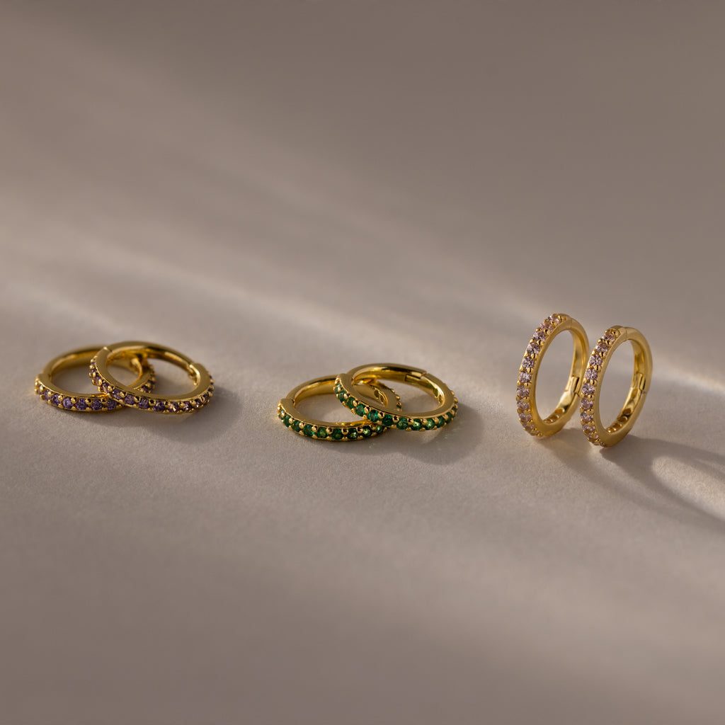 Three pairs of gold hoop earrings with colored gemstones, including Amethyst Clicker Huggies, are displayed on a beige surface under soft lighting.