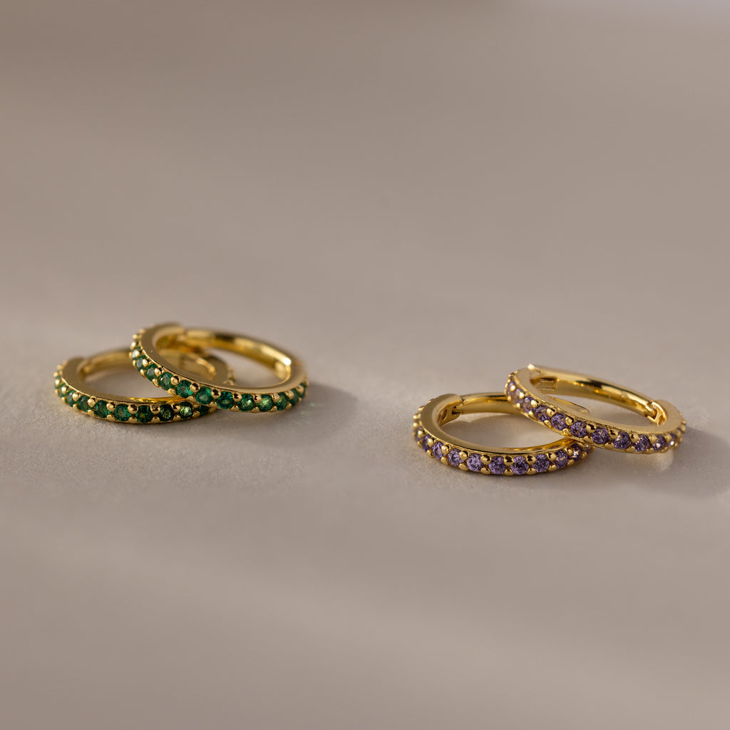 Two pairs of Birthstone Clicker Huggies—one with green stones, the other with purple—rest on a beige surface, making them perfect for everyday wear or as birthstone earrings.