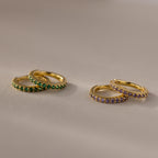 Two pairs of Birthstone Clicker Huggies—one with green stones, the other with purple—rest on a beige surface, making them perfect for everyday wear or as birthstone earrings.